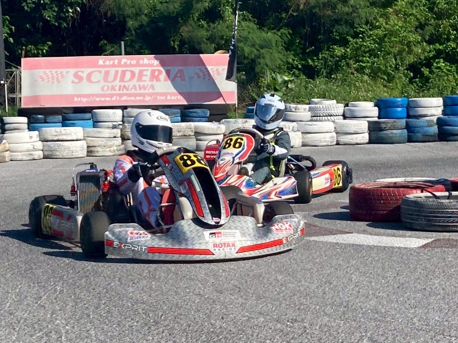 Kart 88 leading kart 86 through a corner past tire stacks and a Scuderia Okinawa banner